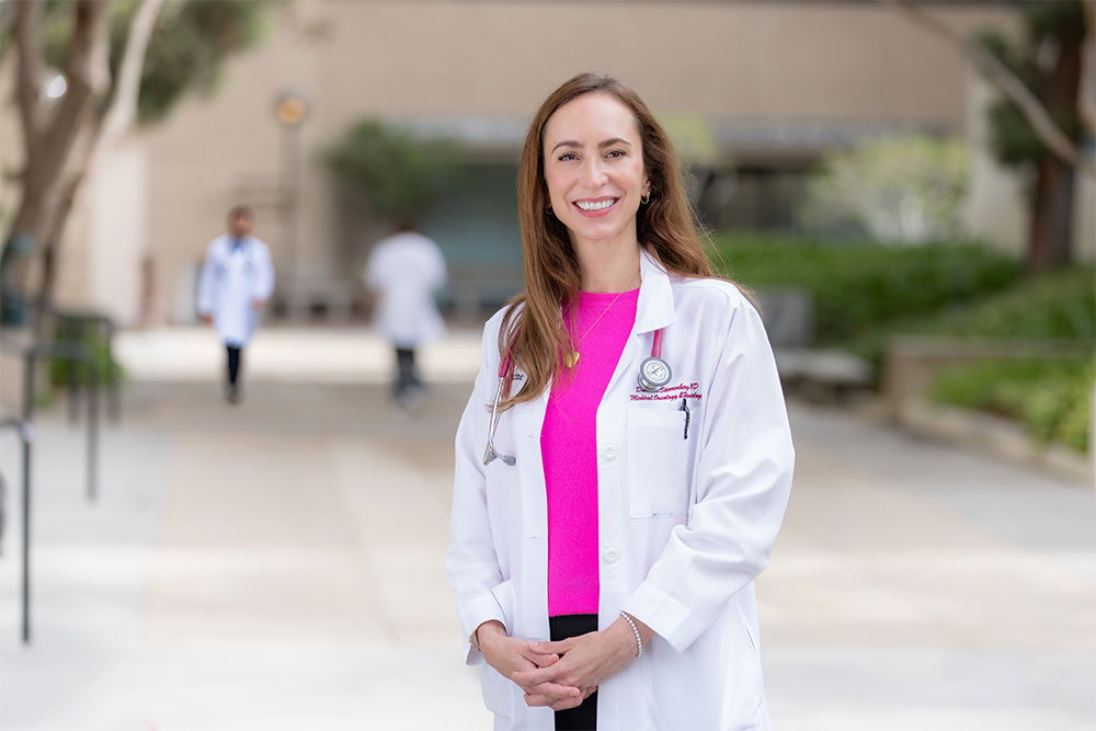 Dr. Danielle Sterrenberg stands outside in a pink shirt and a white medical coat and smiles.